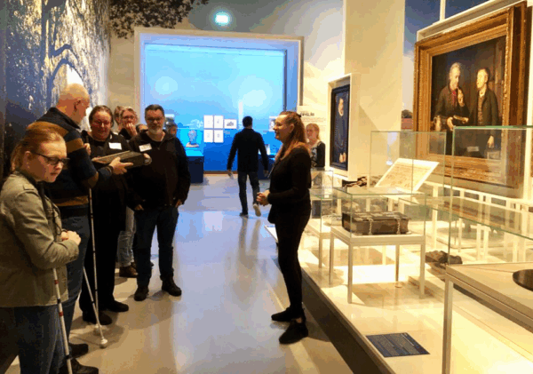 Blinde en slechtziende museumbezoekers luisteren naar de rondleider en betasten een object, vergelijkbaar als in het schilderij waar ze voor staan.