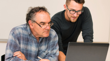 twee mensen kijken naar een beeldscherm; de een zittend, de andere persoon staat en legt iets uit